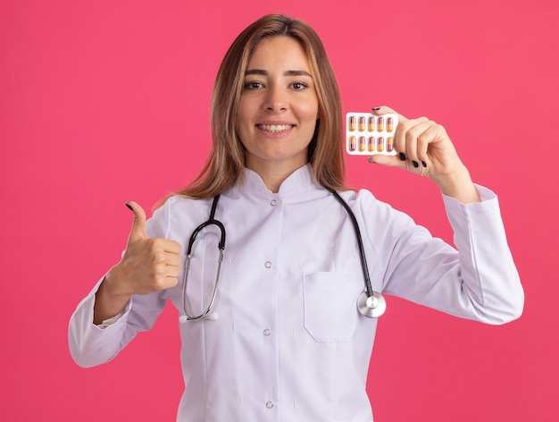 Sonriente joven doctora vistiendo bata médica con estetoscopio sosteniendo píldoras mostrando el pulgar hacia arriba aislado en la pared rosa