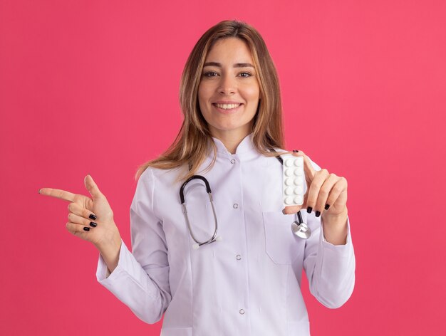 Sonriente joven doctora vistiendo bata médica con estetoscopio sosteniendo píldoras puntos en el lado aislado en la pared rosa