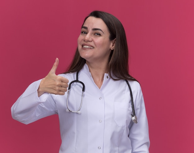 Sonriente joven doctora vistiendo bata médica con estetoscopio Thumbs up en rosa