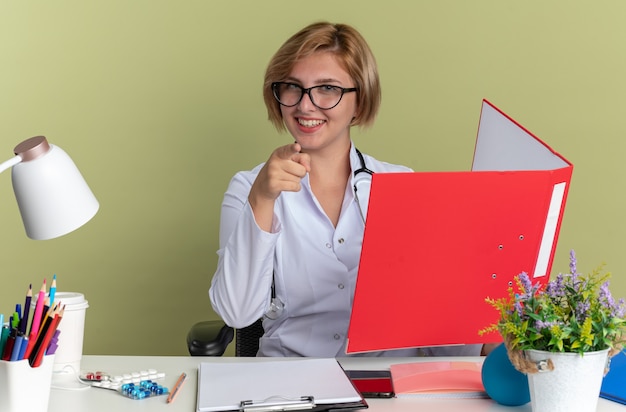 Sonriente joven doctora vistiendo bata médica con gafas y estetoscopio se sienta a la mesa con herramientas médicas sosteniendo carpeta y puntos a cámara aislada sobre fondo verde oliva