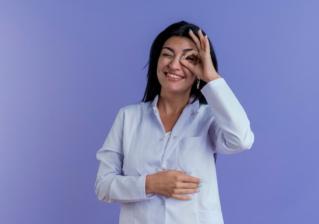 Sonriente joven doctora vistiendo bata médica haciendo gesto de mirada poniendo la mano sobre el vientre aislado en la pared púrpura con espacio de copia