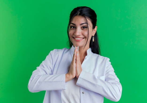 Sonriente joven doctora vistiendo bata médica manteniendo las manos juntas aisladas en la pared verde