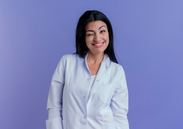 Sonriente joven doctora vistiendo bata médica mirando y guiñando un ojo