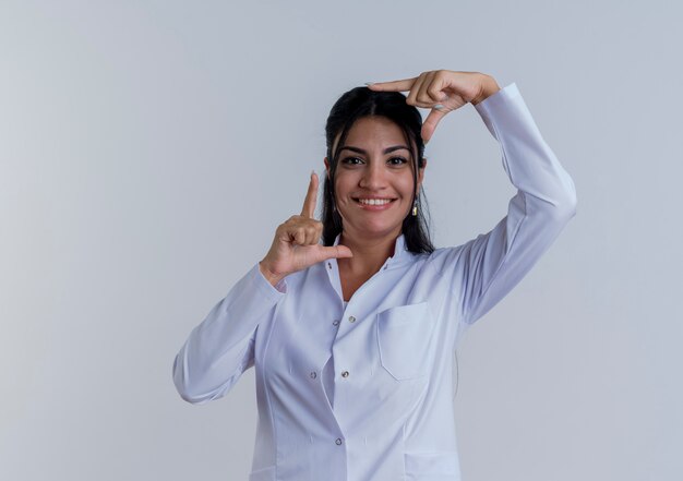 Sonriente joven doctora vistiendo bata médica mirando haciendo gesto de marco aislado