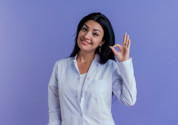 Sonriente joven doctora vistiendo bata médica mirando haciendo signo ok