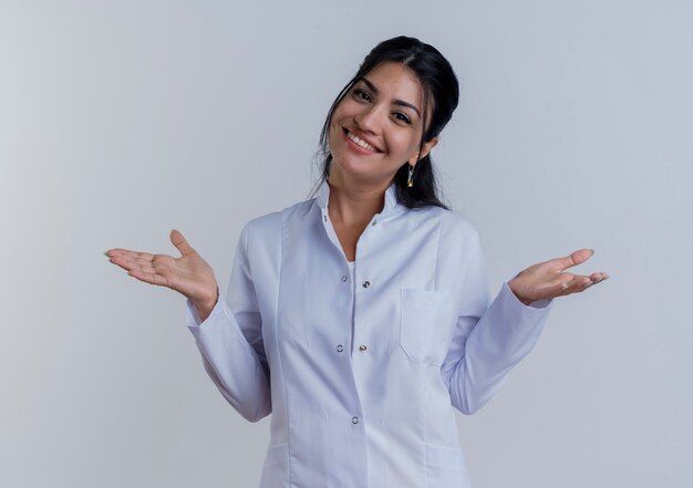 Sonriente joven doctora vistiendo bata médica mirando mostrando las manos vacías aisladas