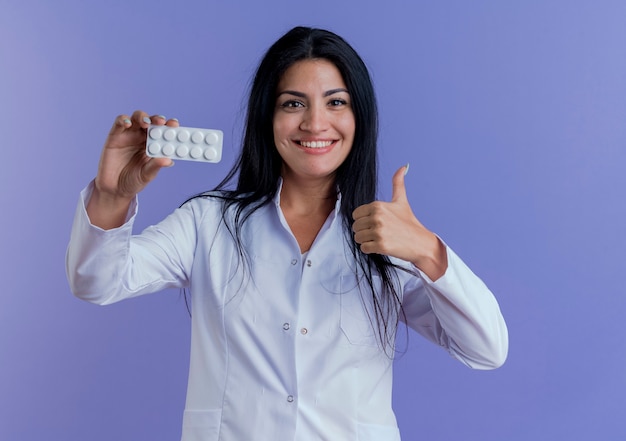 Sonriente joven doctora vistiendo bata médica mostrando el paquete de tabletas médicas, mirando mostrando el pulgar hacia arriba