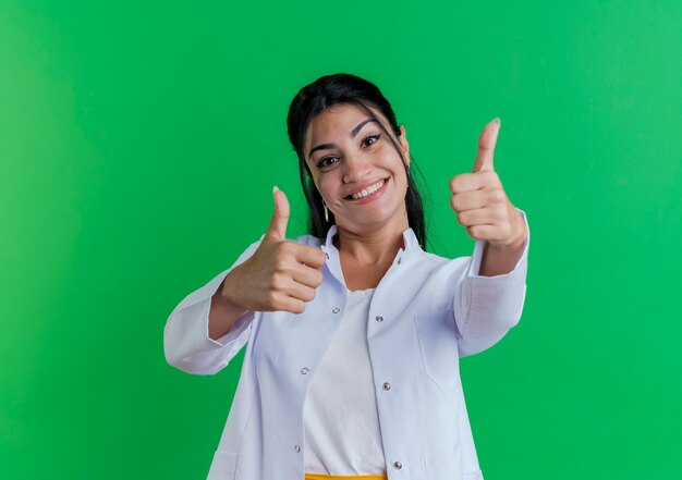 Sonriente joven doctora vistiendo bata médica mostrando los pulgares para arriba aislado en la pared verde con espacio de copia