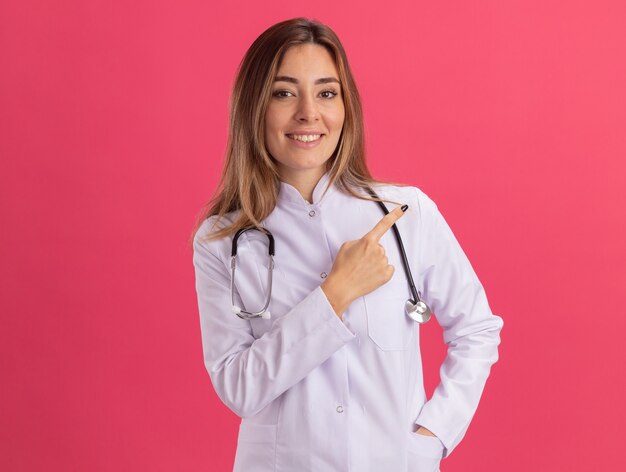 Sonriente joven doctora vistiendo bata médica con puntos de estetoscopio en el lado aislado en la pared rosa con espacio de copia