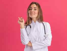 Foto gratuita sonriente joven doctora vistiendo bata médica con puntos de estetoscopio en el lado aislado en la pared rosa con espacio de copia
