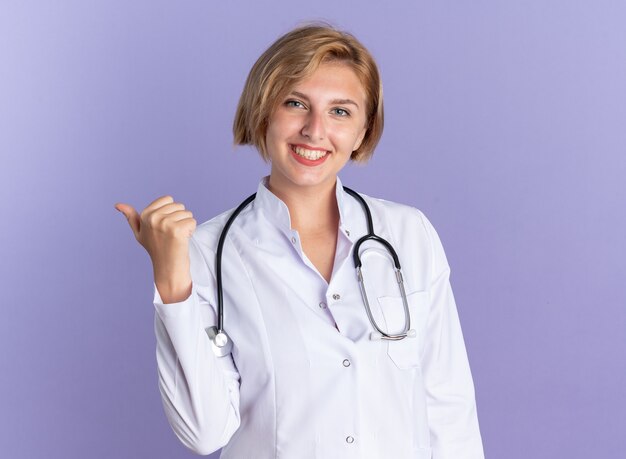 Sonriente joven doctora vistiendo bata médica con puntos de estetoscopio en el lado aislado sobre fondo azul con espacio de copia