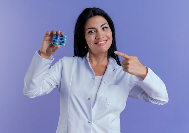 Sonriente joven doctora vistiendo bata médica sosteniendo y apuntando al paquete de cápsulas médicas mirando