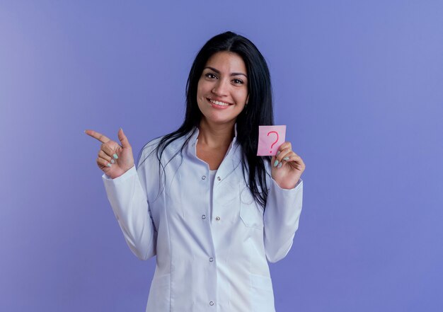 Sonriente joven doctora vistiendo bata médica sosteniendo el signo de interrogación apuntando al lado aislado en la pared púrpura con espacio de copia