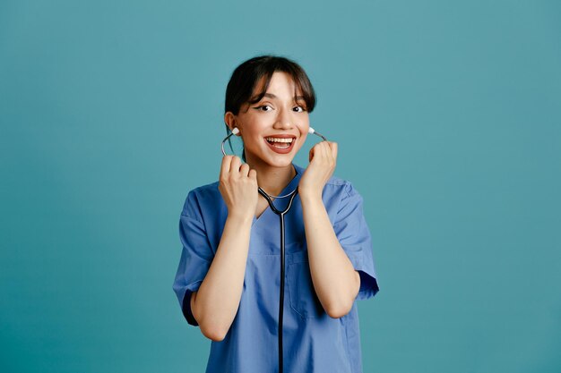 Sonriente joven doctora vistiendo uniforme fith estetoscopio aislado sobre fondo azul.