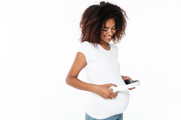 Sonriente joven embarazada mujer africana escuchando música con auriculares.