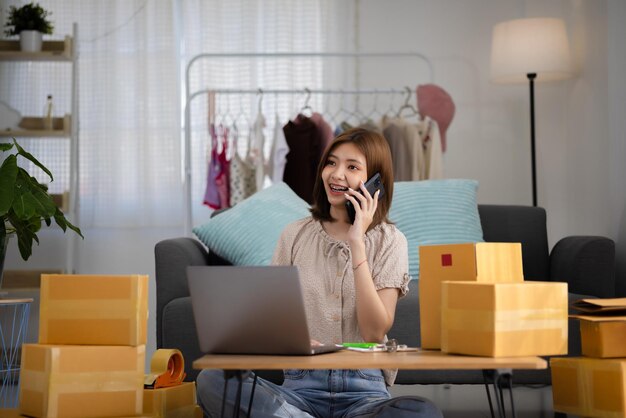 Sonriente joven empresaria asiática feliz que recibe una llamada telefónica para un nuevo pedido de ventas entre cajas de productos con computadora portátil