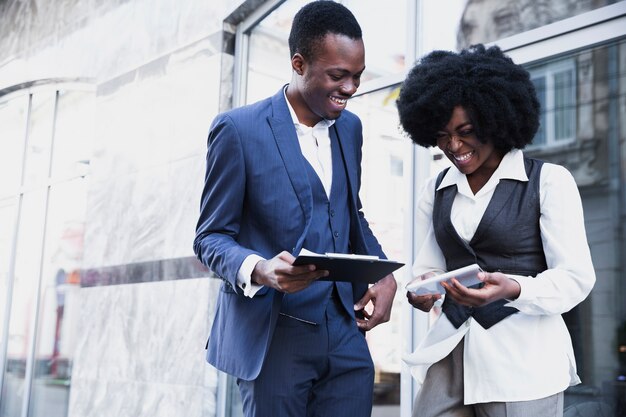 Sonriente joven empresario africano y empresaria sosteniendo portapapeles y tableta digital