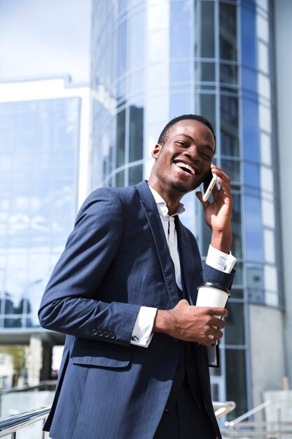 Sonriente joven empresario africano frente a un edificio corporativo hablando por teléfono móvil