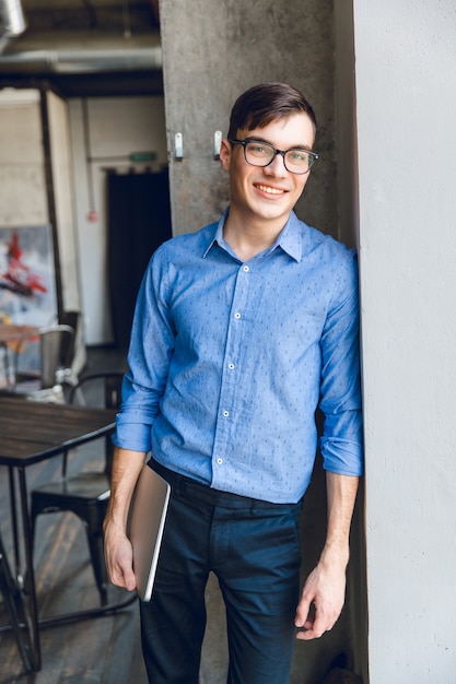 Foto gratuita sonriente joven empresario apoyarse en la pared gris sosteniendo el portátil en la mano