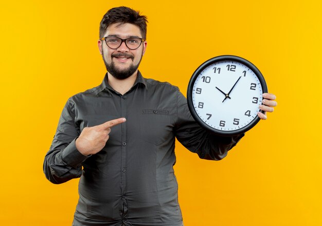 Sonriente joven empresario con gafas sosteniendo y apunta al reloj de pared aislado en amarillo