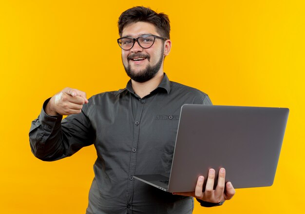 Sonriente joven empresario con gafas sosteniendo portátil y apunta a la cámara