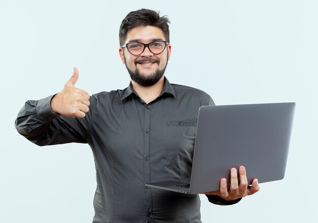 Sonriente joven empresario con gafas sosteniendo portátil su pulgar hacia arriba aislado en blanco