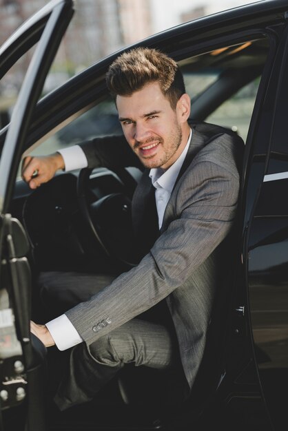 Sonriente joven empresario sentado en el coche con una puerta abierta