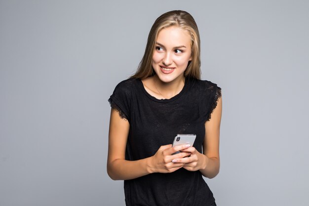 Sonriente joven encantadora de pie y con teléfono celular sobre fondo gris