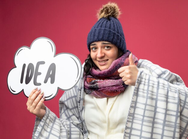 Sonriente joven enferma con bata de invierno sombrero y bufanda envuelta en cuadros sosteniendo burbuja de idea mirando al frente mostrando el pulgar hacia arriba aislado en la pared rosa