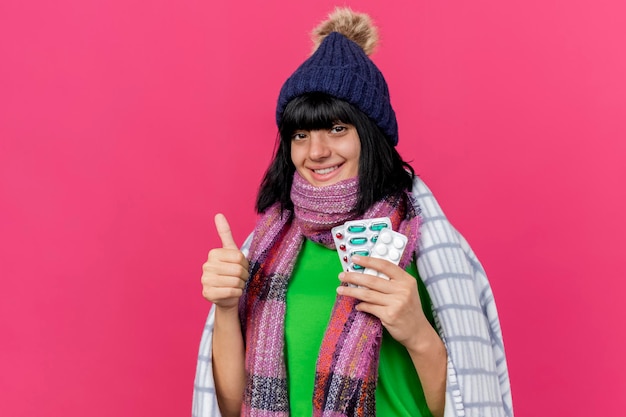 Sonriente joven enferma con gorro de invierno y bufanda envuelta en cuadros con píldoras médicas mirando al frente mostrando el pulgar hacia arriba aislado en la pared rosa con espacio de copia