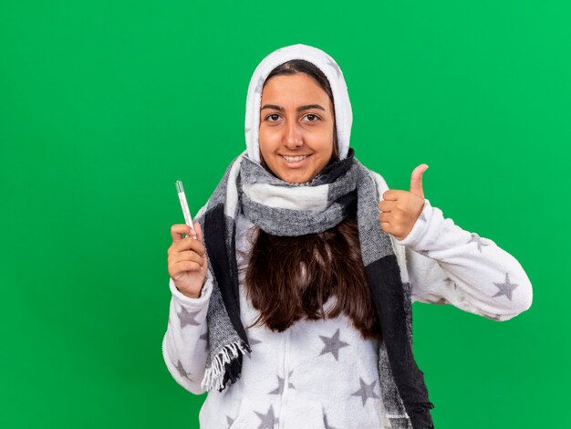 Sonriente joven enferma poniéndose bufanda con capucha vistiendo un termómetro mostrando el pulgar hacia arriba aislado en verde