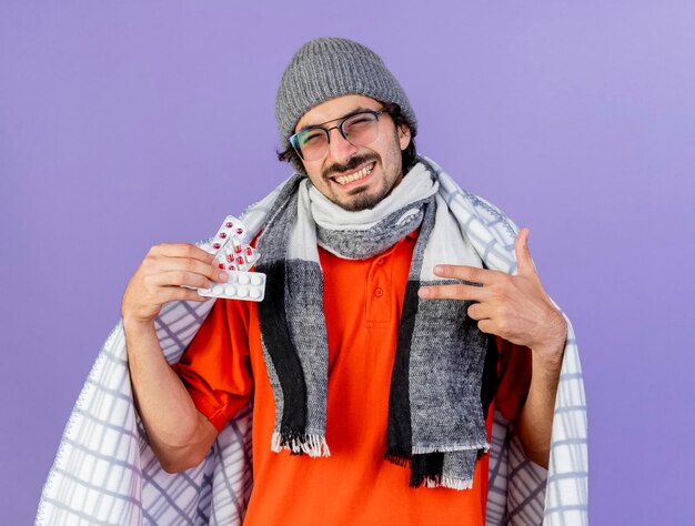 Sonriente joven enfermo con gafas, gorro de invierno y bufanda envuelto en cuadros sosteniendo y apuntando a paquetes de píldoras médicas mirando al frente aislado en la pared púrpura