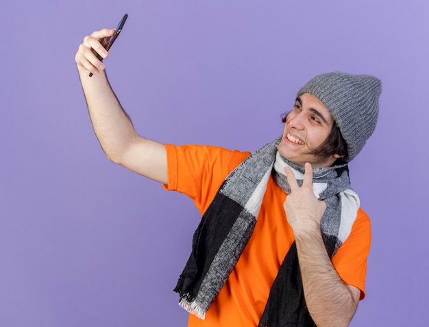 Sonriente joven enfermo con sombrero de invierno con bufanda tomar un selfie mostrando gesto de paz aislado sobre fondo púrpura