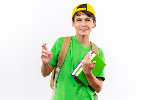 Sonriente joven escolar con mochila con gorra sosteniendo libros cruzando los dedos aislado en la pared blanca