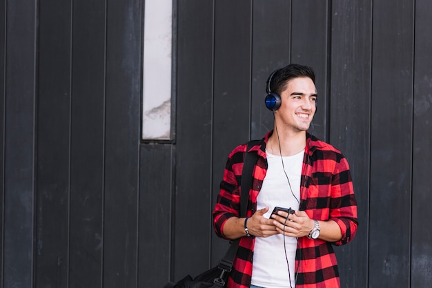 Foto gratuita sonriente joven escuchando música en frente de la pared de madera negra