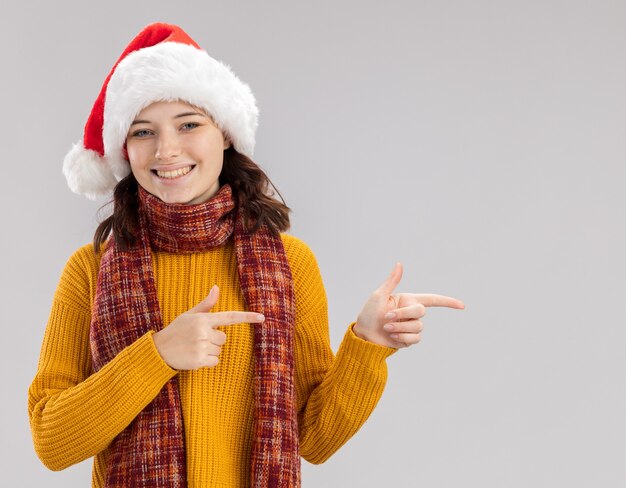 Sonriente joven eslava con gorro de Papá Noel y bufanda alrededor del cuello apuntando al lado aislado en la pared blanca con espacio de copia