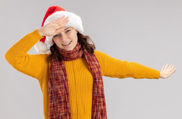 Sonriente joven eslava con gorro de Papá Noel y bufanda alrededor del cuello haciendo dab aislado en la pared blanca con espacio de copia