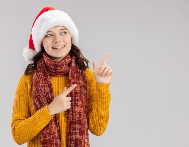 Sonriente joven eslava con gorro de Papá Noel y con bufanda alrededor del cuello mirando y apuntando al lado aislado en la pared blanca con espacio de copia