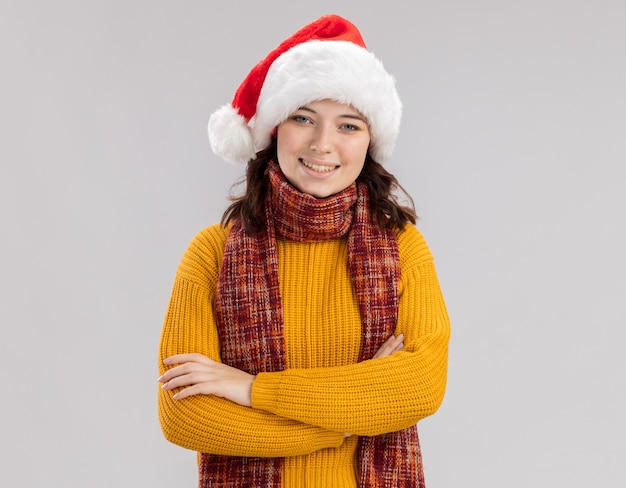 Sonriente joven eslava con gorro de Papá Noel y bufanda alrededor del cuello de pie con los brazos cruzados aislado en la pared blanca con espacio de copia