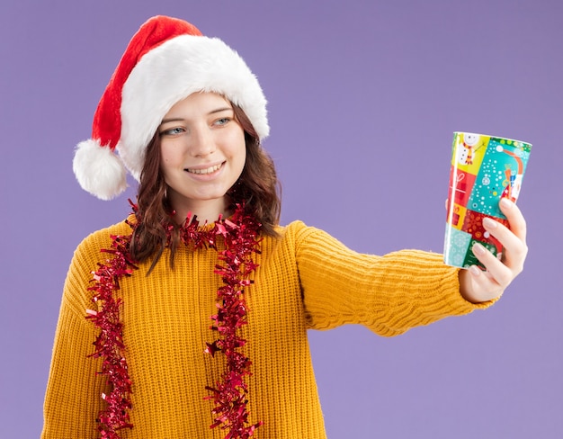 Sonriente joven eslava con gorro de Papá Noel y con guirnalda alrededor del cuello sosteniendo y mirando un vaso de papel aislado sobre fondo púrpura con espacio de copia