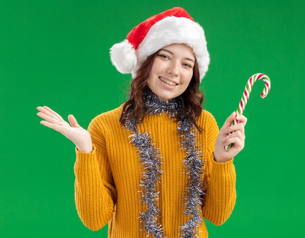 Sonriente joven eslava con gorro de Papá Noel y con guirnalda alrededor del cuello sostiene bastón de caramelo y mantiene la mano abierta