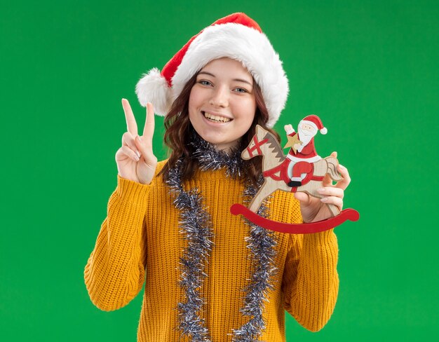 Sonriente joven eslava con sombrero de santa y con guirnalda alrededor del cuello sosteniendo a santa en la decoración del caballo mecedora y gesticulando el signo de la victoria