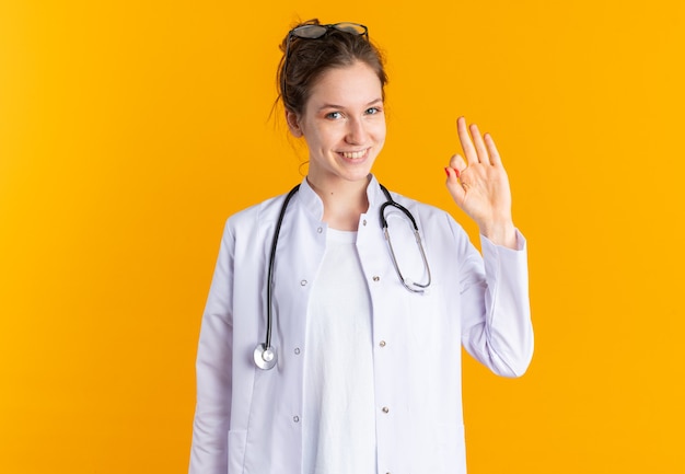 Sonriente joven eslava en uniforme médico con estetoscopio gesticulando signo ok aislado en la pared naranja con espacio de copia