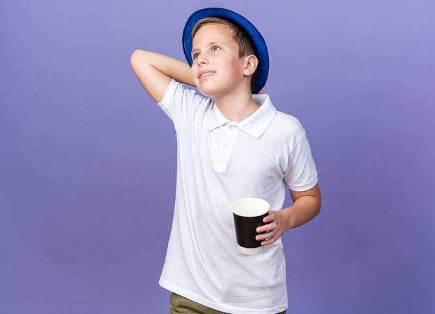 Sonriente joven eslavo con gorro de fiesta azul poniendo la mano en la cabeza detrás y sosteniendo un vaso de papel mirando al lado aislado en la pared púrpura con espacio de copia