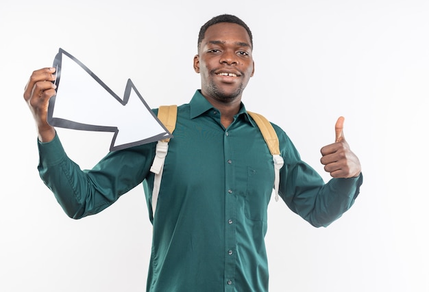 Sonriente joven estudiante afroamericano con mochila sosteniendo la flecha apuntando a un lado y apuntando hacia arriba