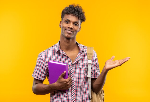 Sonriente joven estudiante afroamericano con mochila sosteniendo el libro y manteniendo la mano abierta