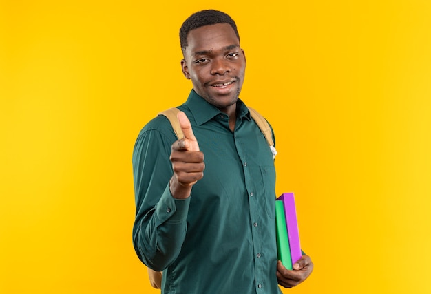 Sonriente joven estudiante afroamericano con mochila sosteniendo libros y apuntando al frente