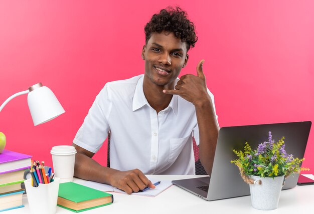 Sonriente joven estudiante afroamericano sentado en un escritorio con herramientas escolares gesticulando llámame señal