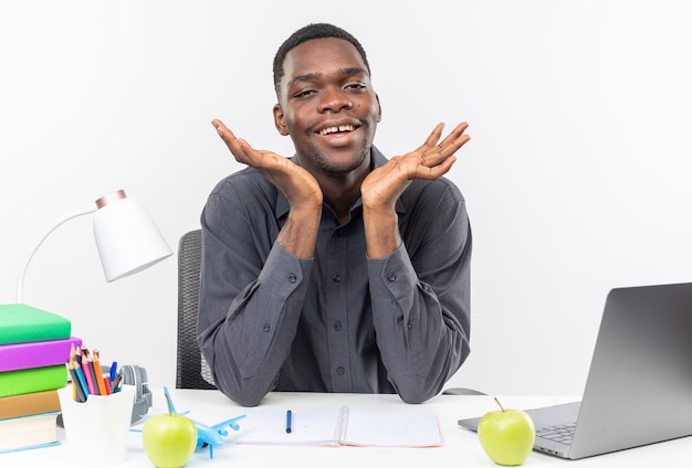 Sonriente joven estudiante afroamericano sentado en el escritorio con herramientas escolares manteniendo las manos abiertas