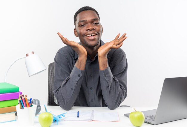 Sonriente joven estudiante afroamericano sentado en el escritorio con herramientas escolares manteniendo las manos abiertas